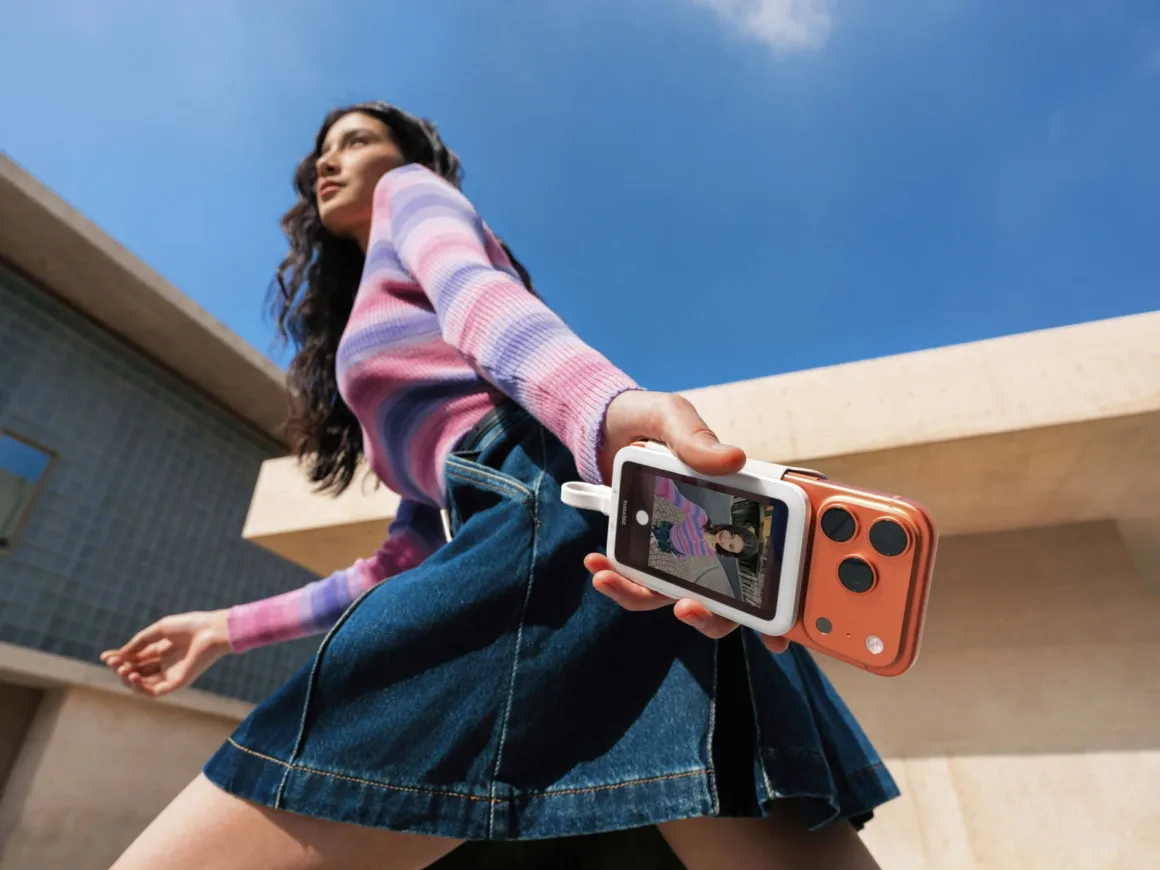 Pembe-mor çizgili kazak ve kot etek giyen bir kadın, elinde anlık fotoğraf makinesi ve basılmış fotoğrafla açık havada yürüyor. Arka planda bina ve mavi gökyüzü var.