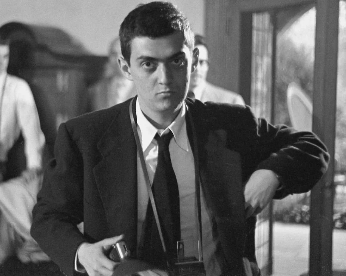 Takım elbise ve kravat giymiş genç bir adam, elinde kamera tutarak doğrudan izleyiciye bakıyor. Arka planda doğal ışıkla aydınlatılmış, hafif flu bir iç mekân ve insanlar görülüyor.