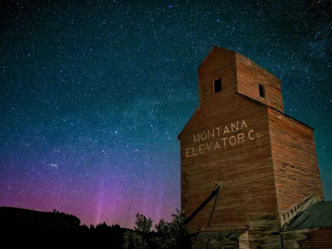 “Montana Elevator Co.” yazılı eski bir ahşap tahıl silosu, ufukta hafif mor bir aurora ile birlikte berrak ve yıldızlarla dolu bir gece gökyüzünün altında duruyor.
