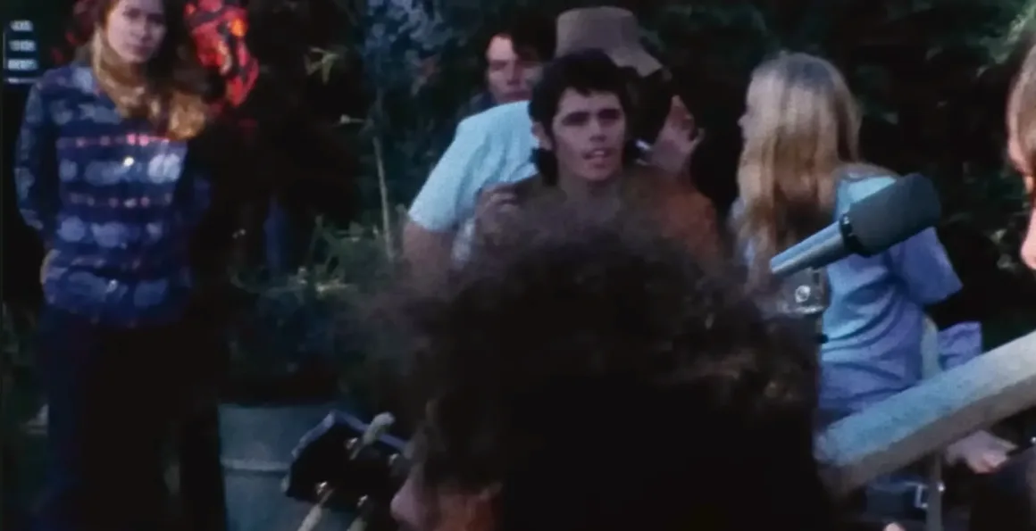 Açık alanda bir grup insan, bazıları sohbet ediyor bazıları dinliyor, ortadaki kişi kameraya doğru bakıyor, çevrede ekipman ve bitkiler görülüyor.