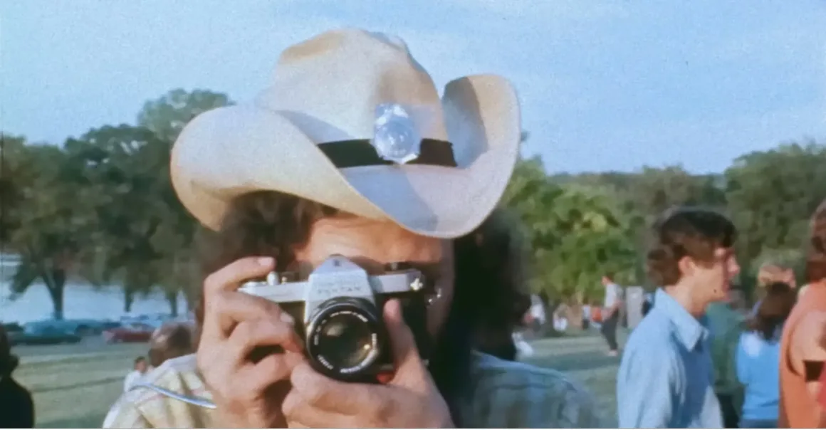 Açık hava etkinliğinde kovboy şapkası ve gözlük takan bir kişi film kamerasıyla fotoğraf çekiyor, arka planda kalabalık ve ağaçlar yer alıyor.
