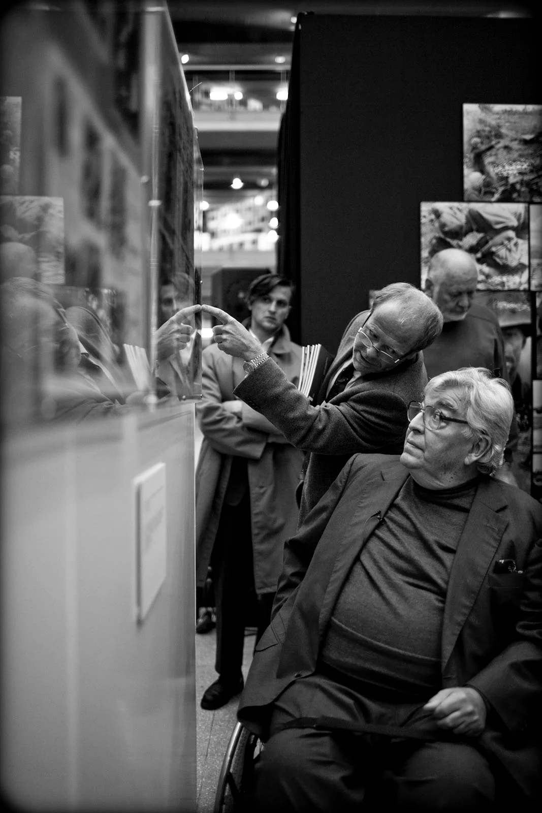 Tekerlekli sandalyedeki bir adam cam arkasındaki bir sergiye bakarken, yanındaki başka bir adam işaret ediyor. Arka planda üç kişi daha sergiyi izliyor. Görüntü siyah beyaz.
