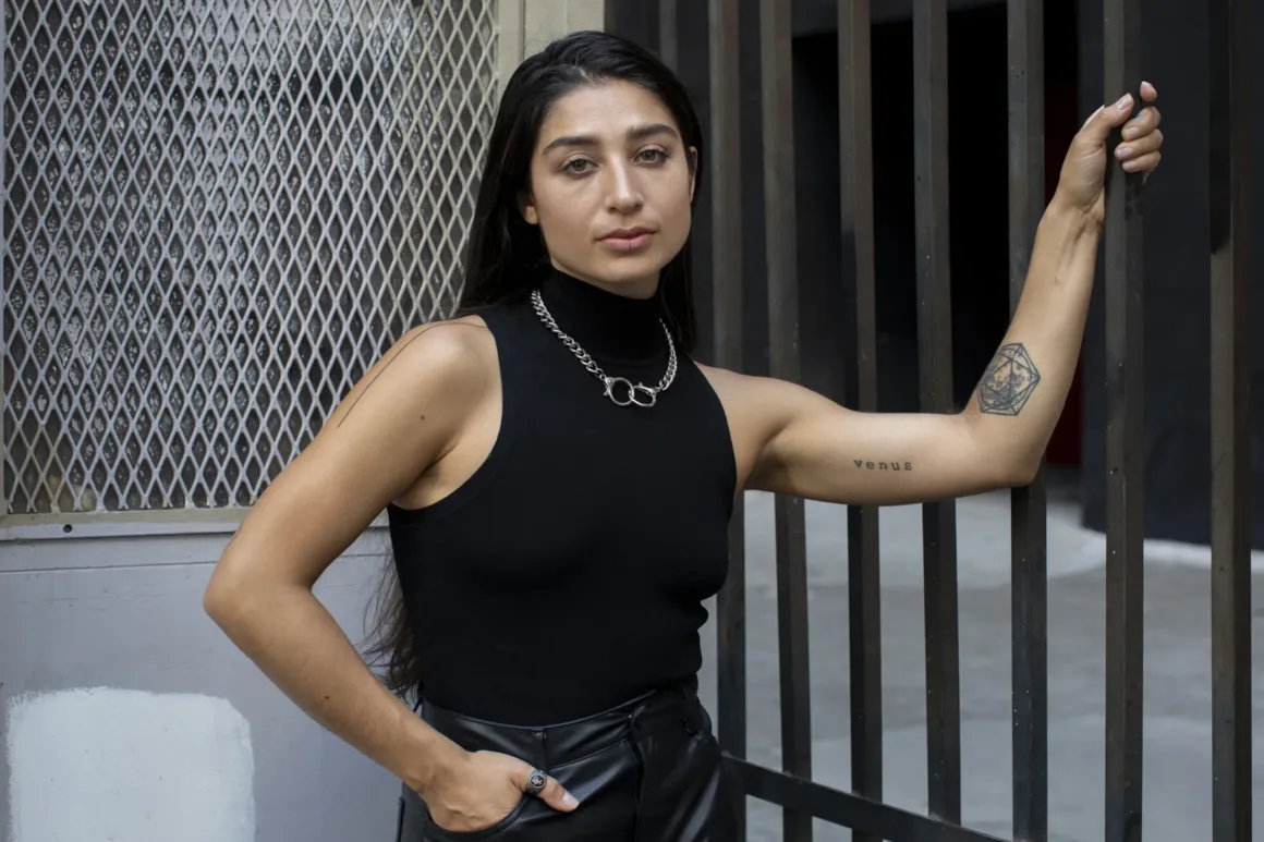 Uzun koyu saçlı, siyah kolsuz boğazlı üst ve zincir kolye takan bir kadın, metal bir kapının yanında duruyor. Bir eli cebinde, diğer eli parmaklıklara dayanmış. Kolunda bir dövme var.