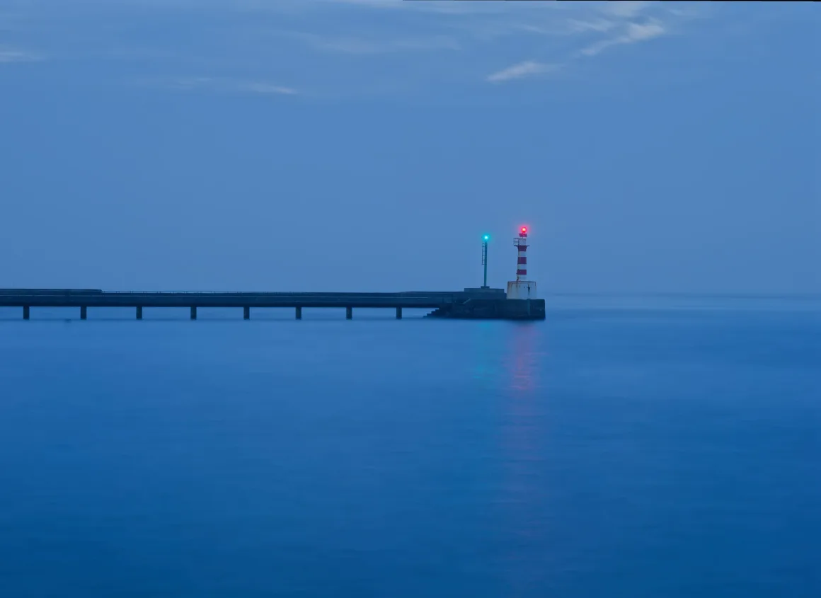 Fotoğrafçılar İçin Tasarım Neden Vazgeçilmezdir? 12 Gün batımına yakın saatlerde, uzun bir iskelenin sonunda kırmızı ve yeşil ışıklı bir deniz feneri; etrafında sakin mavi su ve bulutlu bir gökyüzü.