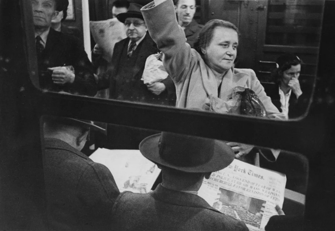 Siyah-beyaz bir metro fotoğrafı: bazı insanlar oturup gazete okurken, bazıları ayakta tutamaklara tutunuyor. Ortada bir kadın kolunu kaldırmış şekilde ayakta duruyor ve sakin bir ifadeyle ileriye bakıyor.