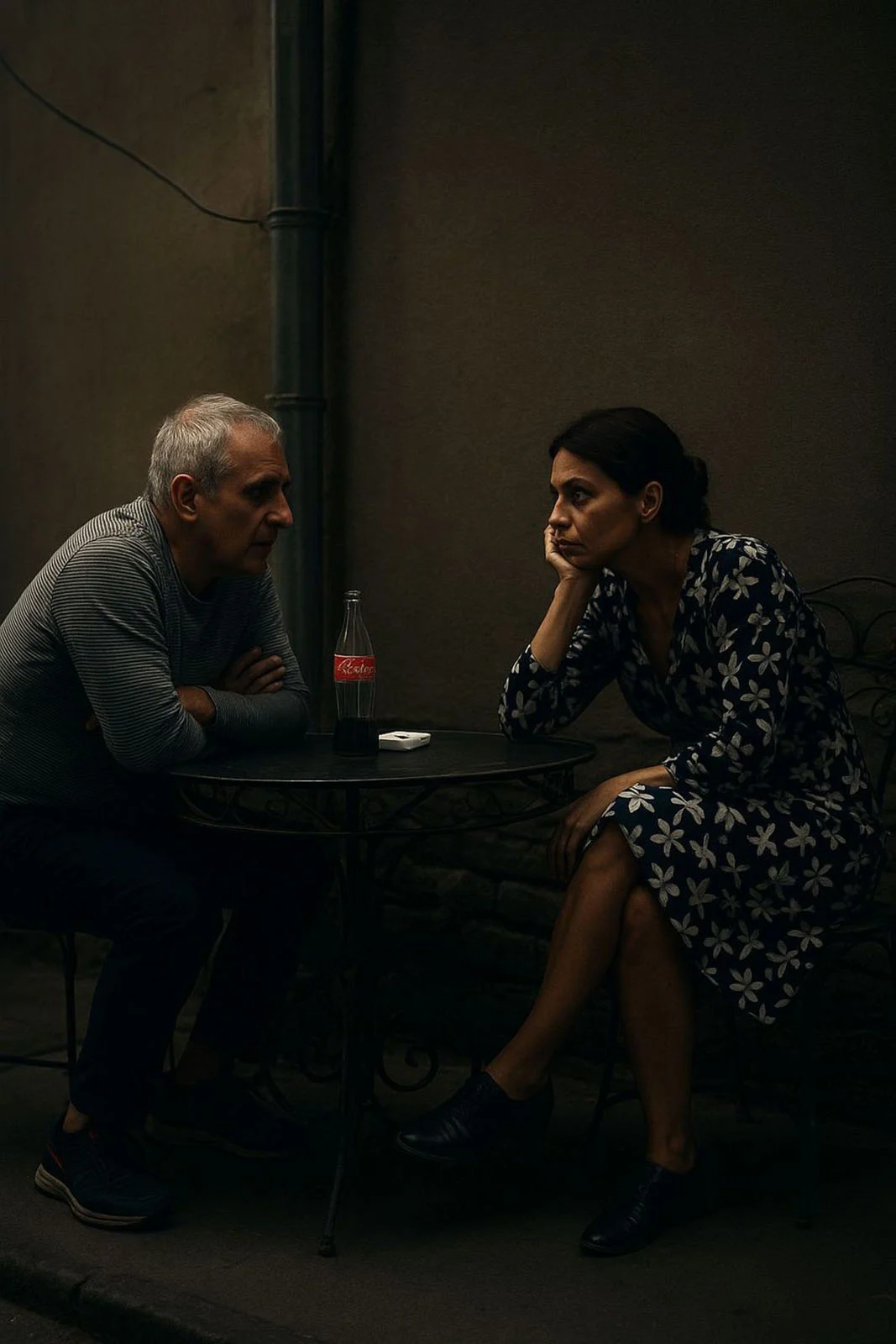 Açık havada küçük yuvarlak bir masada oturan bir kadın ve erkek ciddi bir sohbet içinde; masada Coca-Cola şişesi ve sigara paketi bulunuyor.