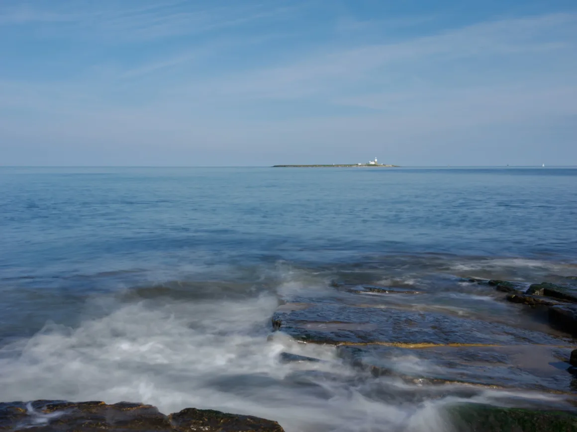 Uzakta küçük bir ada üzerindeki deniz feneri mavi gökyüzü altında görülüyor. Ön planda ise dalgalar koyu kayalık kıyıya hafifçe vuruyor.