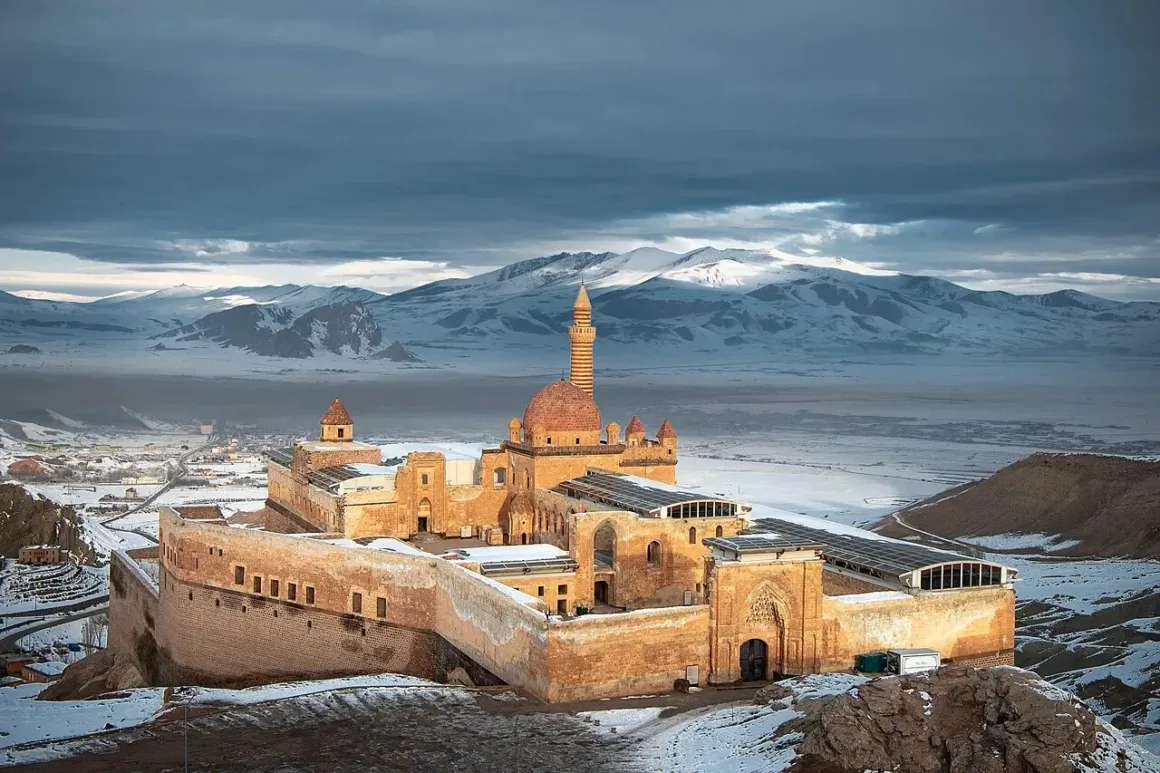 Bulutlu gökyüzü altında, yumuşak gün ışığıyla aydınlanan geniş bir vadide ve karla kaplı dağların arasında yer alan, kubbeli çatılara ve yüksek bir minareye sahip büyük tarihi taş saray.