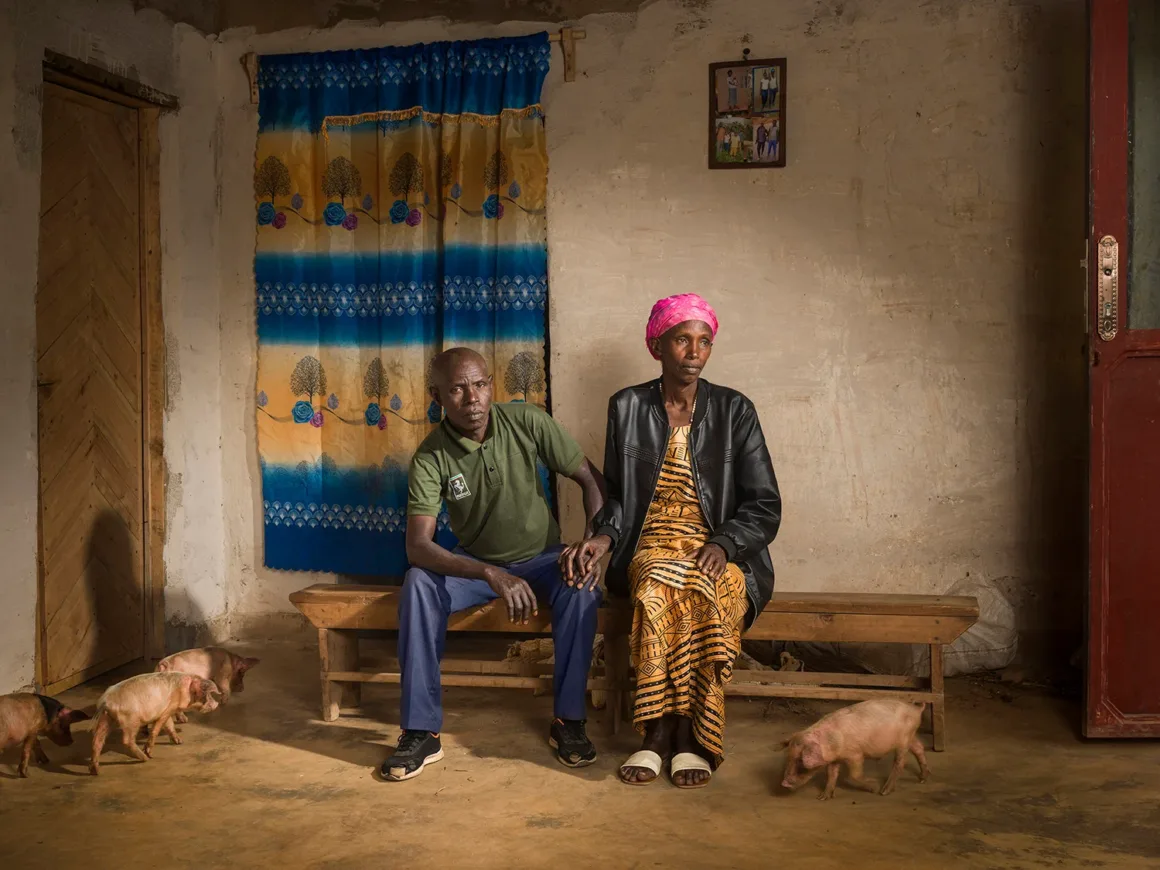 Blood Bonds: Ruanda Soykırımı Sonrası Uzlaşma ve Affetmenin Fotoğrafları
Mavi ve sarı bir perdenin bulunduğu, duvarda bir fotoğrafın asılı olduğu sade bir odada, ahşap bir bankta oturan bir kadın ve bir erkek. Yerde üç yavru domuz bulunuyor. Kadın pembe bir başörtüsü ve desenli bir elbise giyiyor; erkek gündelik kıyafetler içinde.
