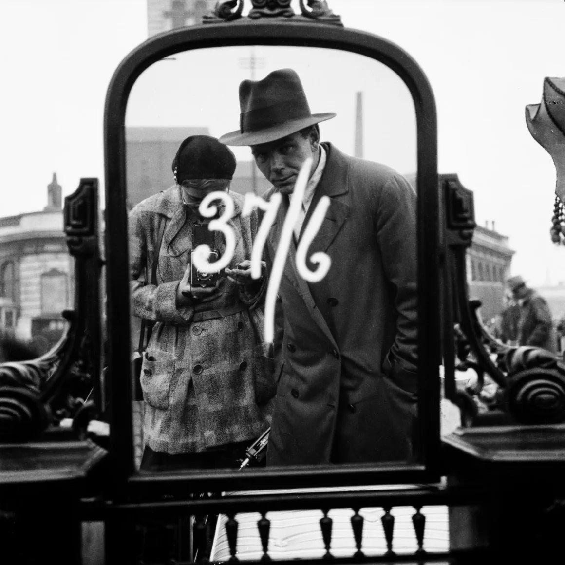 Bir Fotoğrafçı, İki Hayat: Sovyet Ajanına Dönüşen İngiliz Kadının Gizli Tarihi
Siyah-beyaz bir fotoğrafta, “37 1/6” yazılı süslü bir aynada yansıyan palto ve şapka giymiş iki kişi görülüyor. Sahne, arka planda binaların yer aldığı bir pazar alanında, açık havada geçiyor gibi görünüyor.