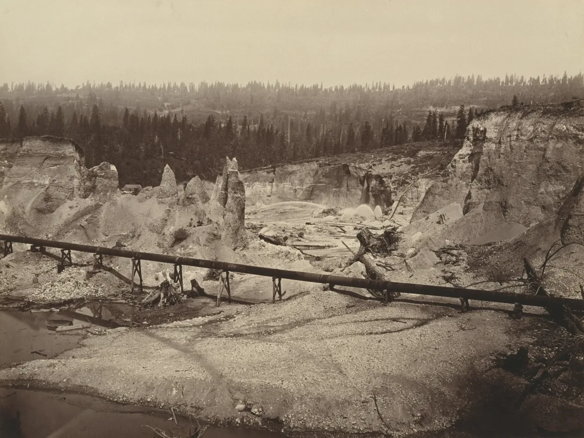200 Yıllık Fotoğraflar Amerika’nın Madencilik Tarihini Gösteriyor 6 Sepya tonlarında bir fotoğrafta aşınmış kayalıklarla dolu çıplak bir arazi ve sahne boyunca uzanan uzun ahşap bir boru hattı görülüyor. Arka planda seyrek ağaçlar ve ormanlık tepeler yer alıyor.
