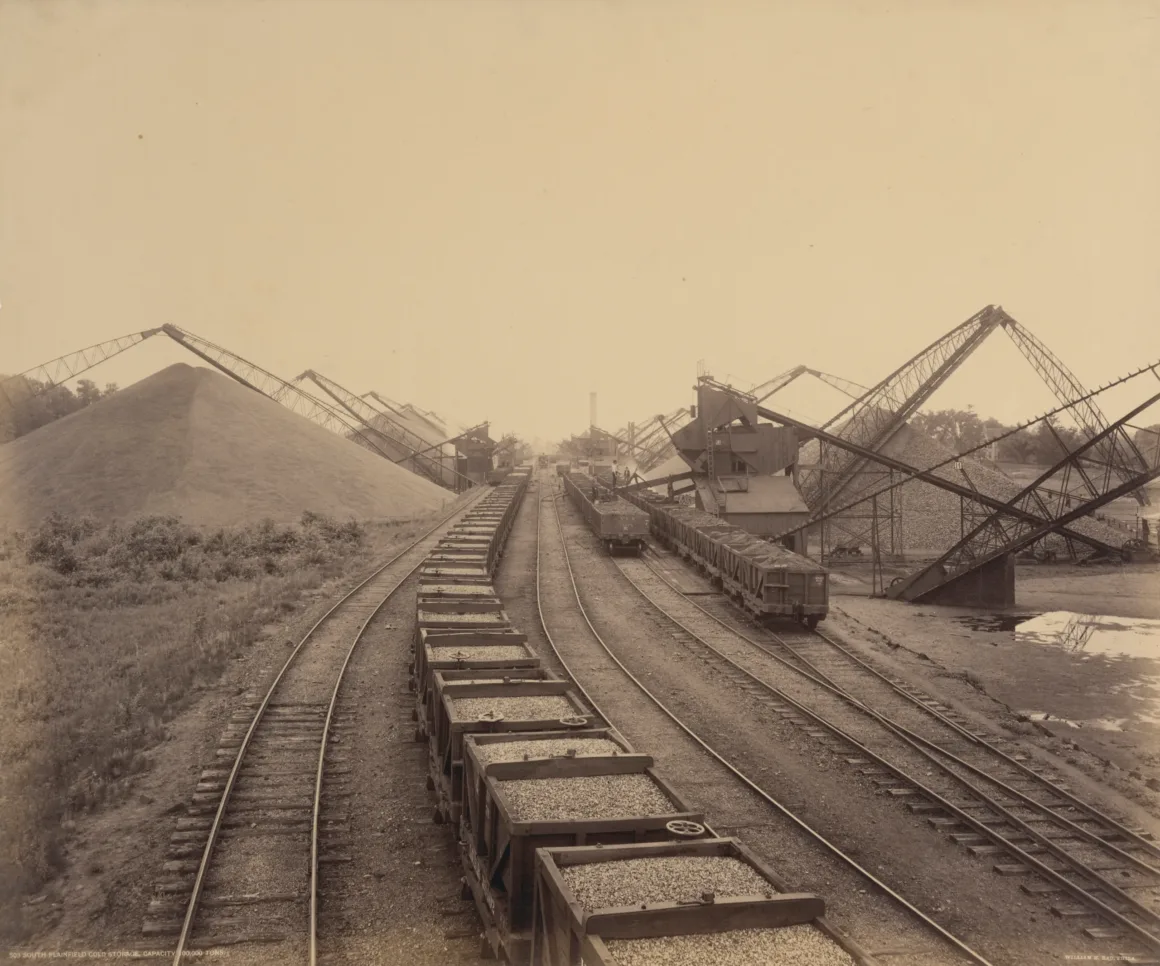 200 Yıllık Fotoğraflar Amerika’nın Madencilik Tarihini Gösteriyor 9 Sepya tonlu fotoğrafta çakıl veya kömür taşıyan trenlerin bulunduğu çok sayıda demiryolu hattı, büyük malzeme yığınları ve rayların üzerinde duran endüstriyel vinçler görülüyor. Arka planda ağaçlar ve bir nehir yer alıyor.