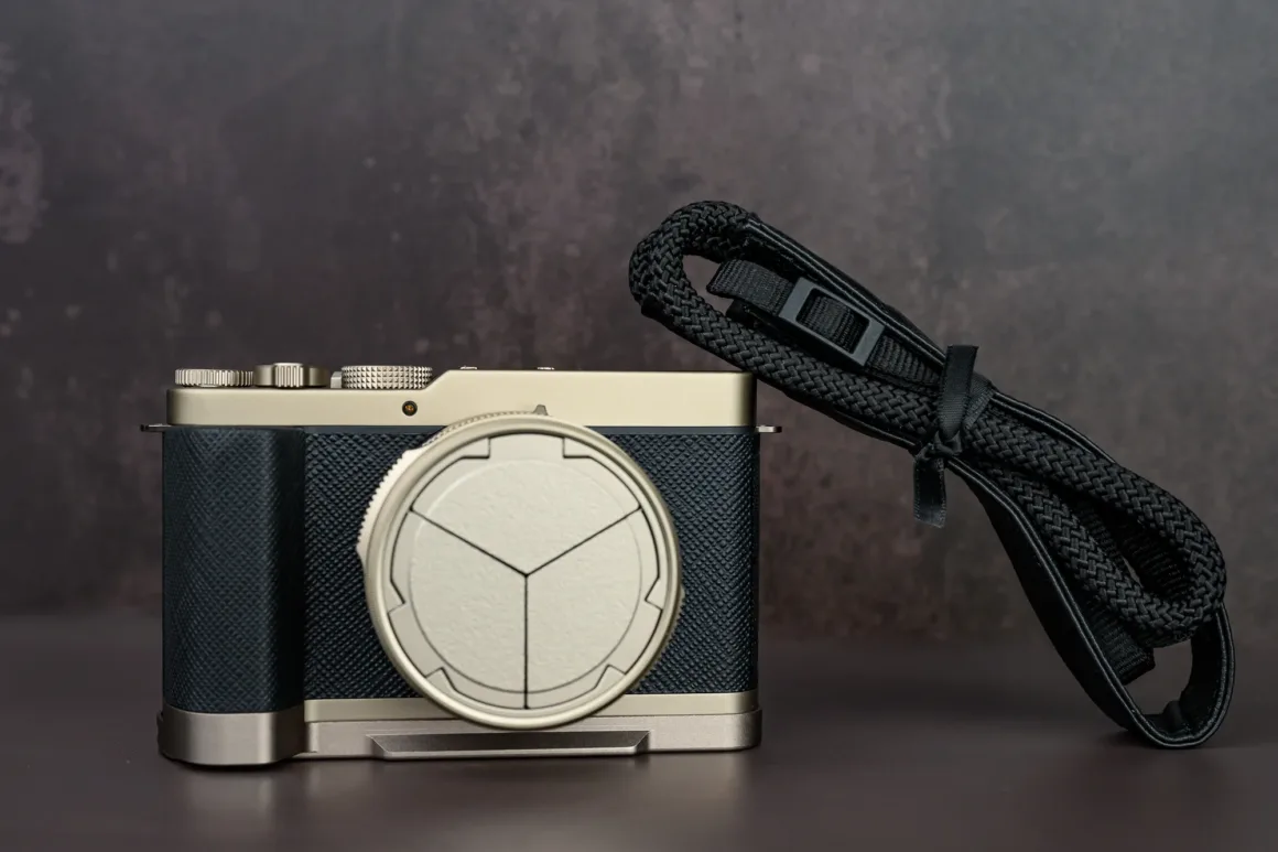 Panasonic Lumix L10 İncelemesi: Kompakt Kameraların Geri Dönüşü 4 Dokulu siyah ve gümüş gövdeye sahip modern dijital kamera, geometrik desenli lens kapağıyla birlikte bir yüzey üzerinde duruyor. Yanında siyah kamera kayışı yer alıyor.