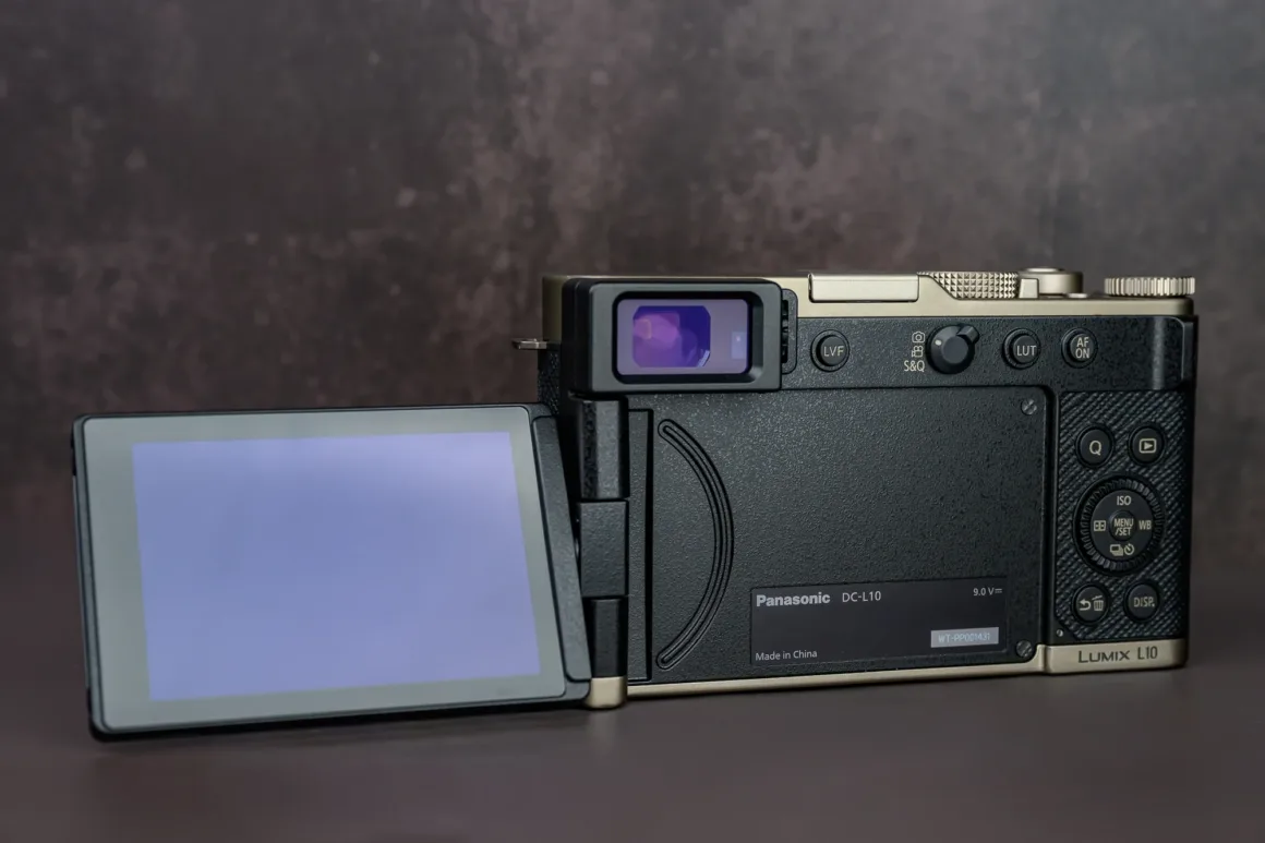 Panasonic Lumix L10 İncelemesi: Kompakt Kameraların Geri Dönüşü 13 Arkadan görünen dijital kameranın yana açılmış LCD ekranı dikkat çekiyor. Sağ tarafta çeşitli kontrol düğmeleri ve elektronik vizör bulunuyor. Kamera kahverengi bir yüzey üzerinde duruyor.