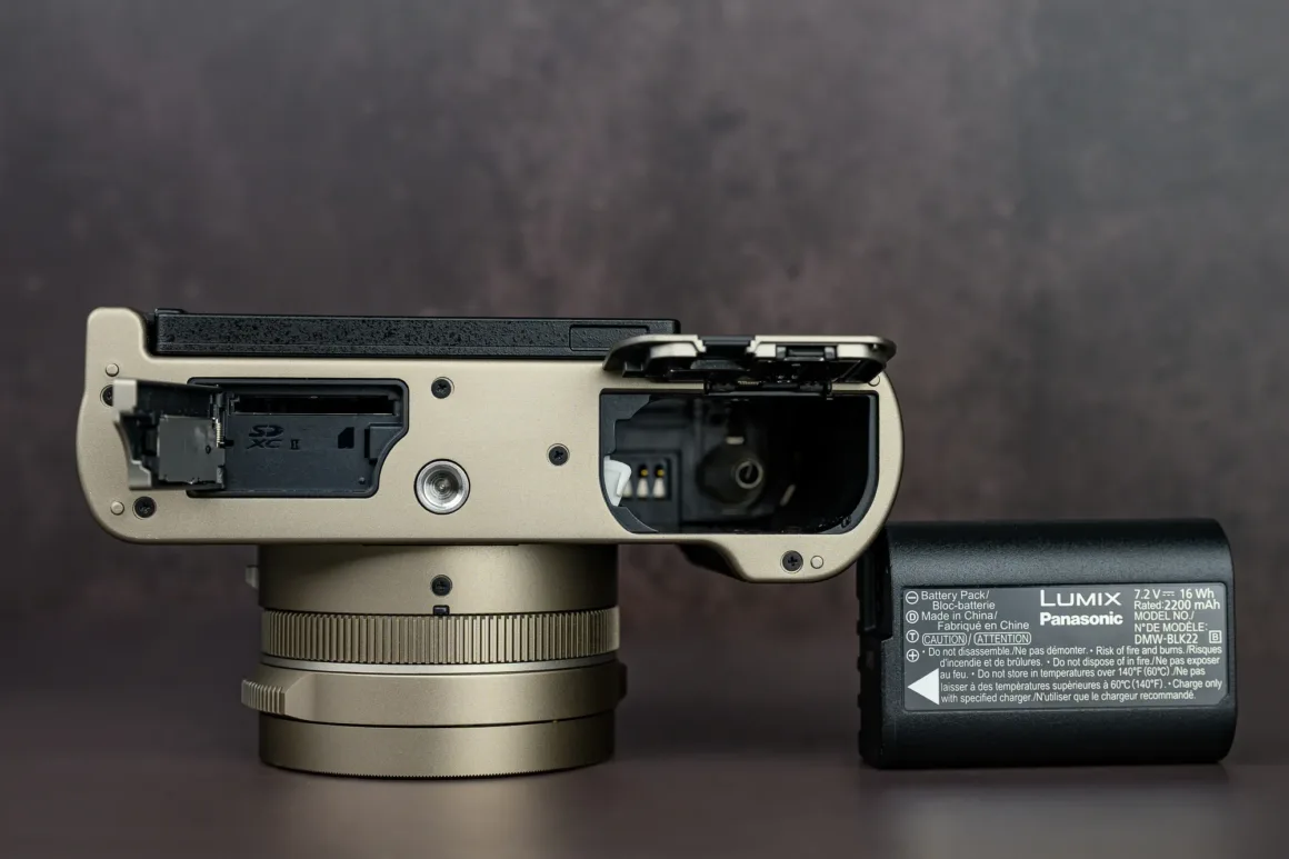 Panasonic Lumix L10 İncelemesi: Kompakt Kameraların Geri Dönüşü 6 Alt kısmı açık dijital kamera görüntüsü. SD kart ve batarya bölmeleri açılmış durumda ve batarya çıkarılmış. Gövde ile batarya karanlık ve bulanık bir yüzey üzerinde duruyor.