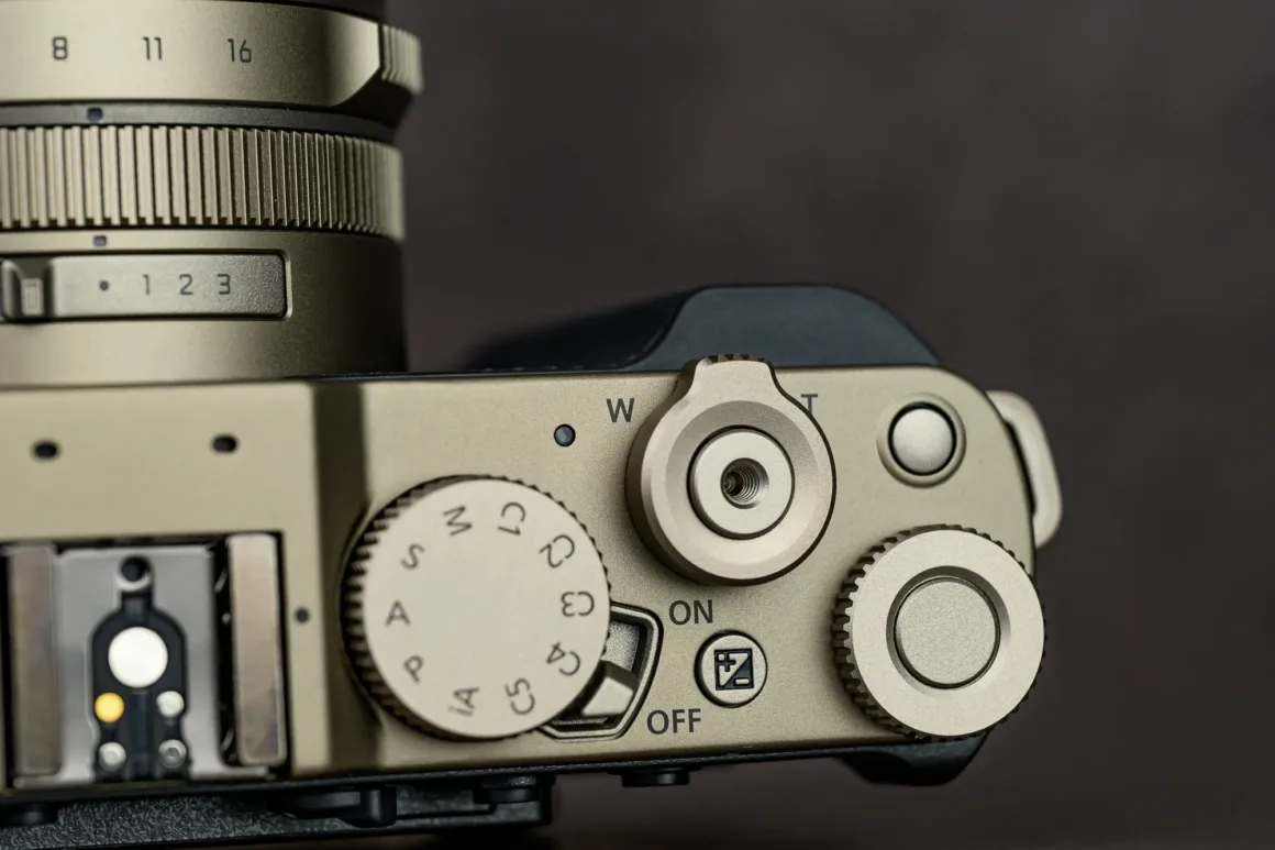 Panasonic Lumix L10 İncelemesi: Kompakt Kameraların Geri Dönüşü 9 Metalik kameranın üst kontrollerinin yakın plan görüntüsü. Ayar kadranları, mod tekeri, açma kapama anahtarı, "C" yazılı düğme ve lens üzerindeki netleme ile zoom işaretleri görülüyor.