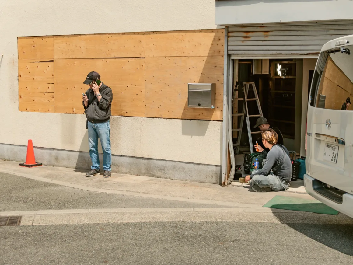 Panasonic Lumix L10 İncelemesi: Kompakt Kameraların Geri Dönüşü 8 Bir adam duvar kenarında telefonla konuşurken, iki kişi yarı açık bir garajın içinde yerde oturuyor. Çevrede araç gereçler ve merdiven bulunuyor. Yakında beyaz bir minibüs park halinde.