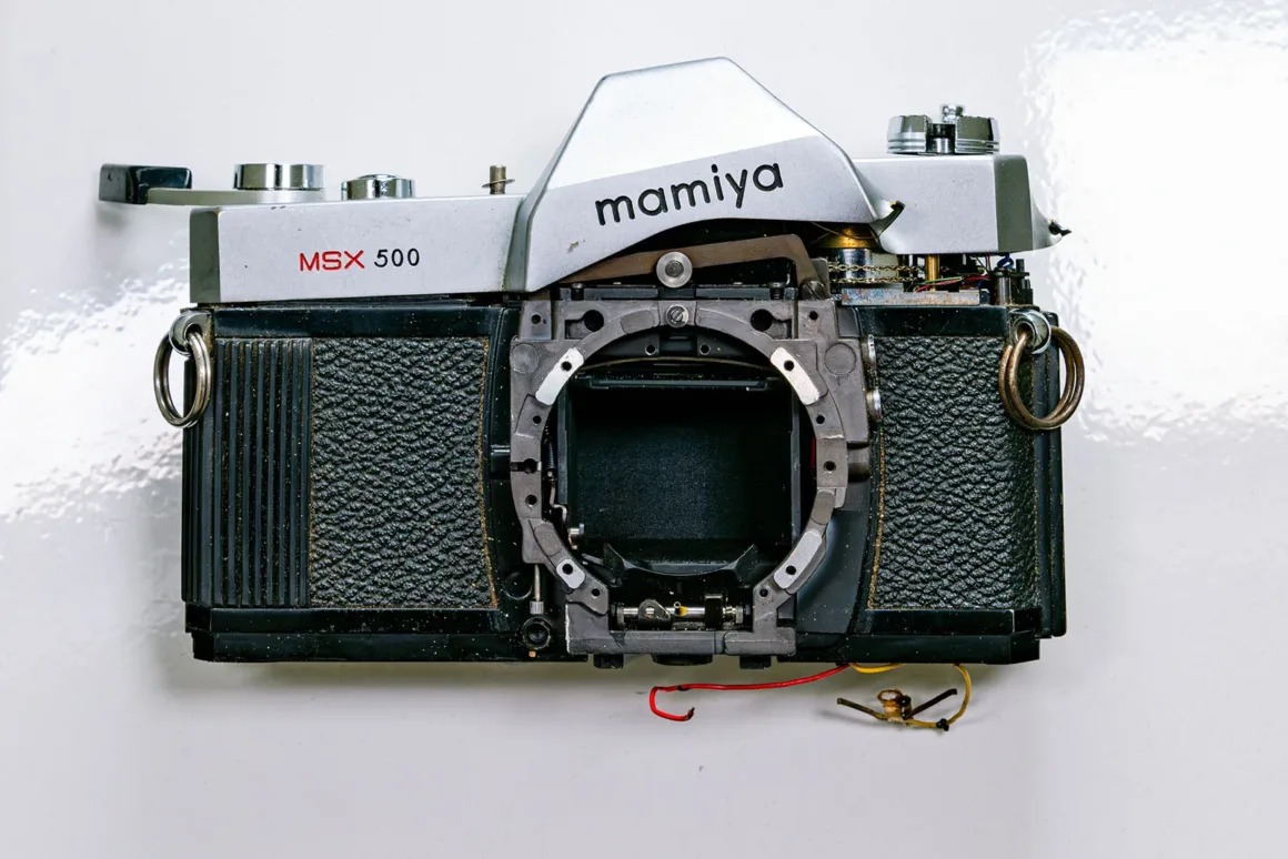 Lensi çıkarılmış ve iç mekanik parçaları görünen parçalarına ayrılmış vintage bir Mamiya MSX 500 film kamerası beyaz yüzey üzerinde duruyor.
