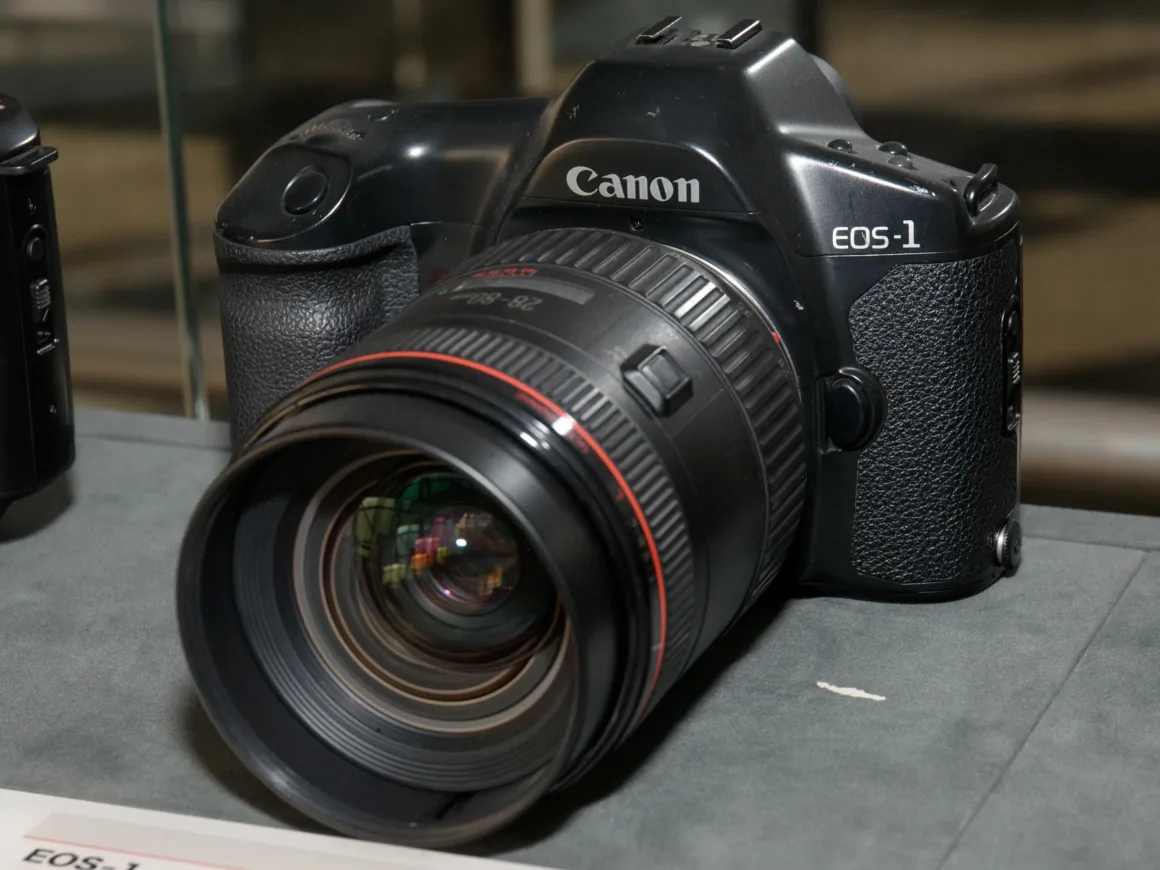 Büyük bir lens takılı Canon EOS-1 kamerası düz bir yüzey üzerinde önden açıyla sergileniyor. Siyah gövde üzerinde marka detayları görünür durumda.