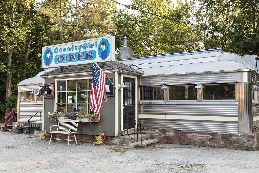 “Country Girl Diner” adlı retro tarzda küçük bir restoran; mavi tabelası, dışarıda Amerikan bayrağı, metal kaplaması ve girişte çiçekler bulunuyor; etrafı ağaçlarla çevrili.