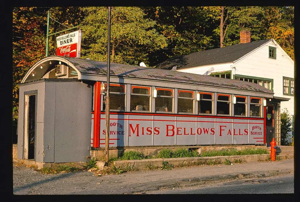 “Miss Bellows Falls” adlı vintage diner; kırmızı beyaz tabelalar, geniş pencereler ve Coca-Cola logosu bulunuyor. Sokak köşesinde, ağaçlar ve bir ev ile çevrili.