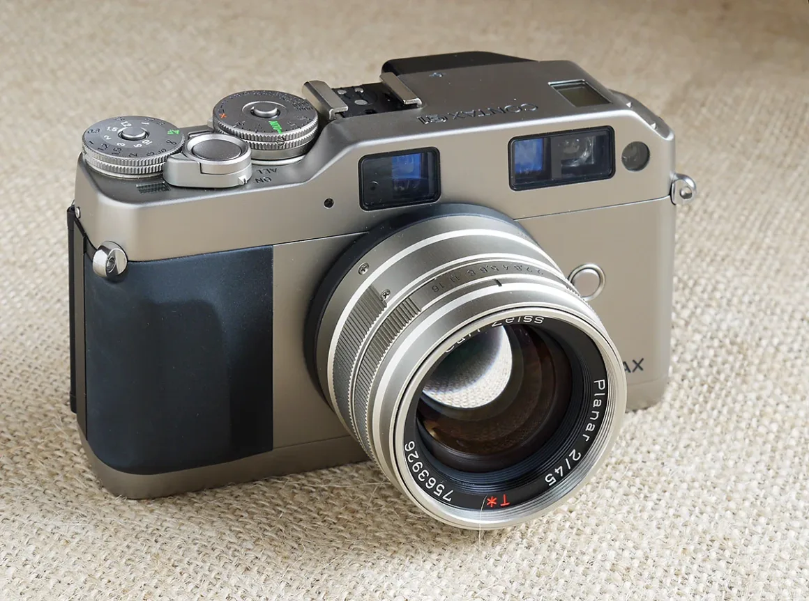 Bej dokulu bir kumaş üzerinde duran, büyük Zeiss Planar 45mm lens takılı gümüş ve siyah renkli Contax G2 film kamerası.