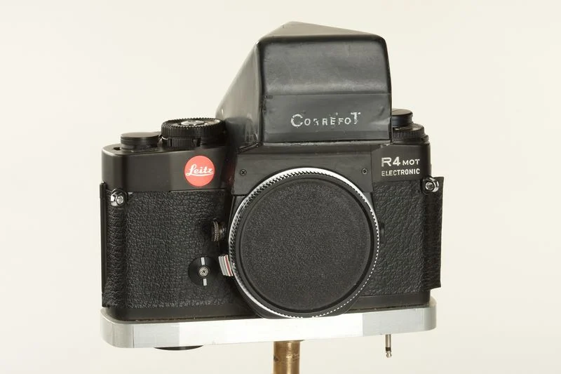 Sade bir arka plan önünde metal stand üzerinde sergilenen, lenssiz siyah gövdeli ve dokulu tutma alanına sahip vintage Cosrefot R4 MOT Electronic prototip 35mm kamera.