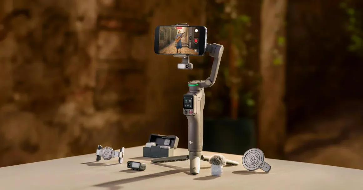 DJI elde taşınabilir gimbal üzerine yerleştirilmiş bir akıllı telefon, çeşitli kamera aksesuarlarının bulunduğu bir masada konumlandırılmış halde görülüyor. Arka plan bulanık açık hava manzarasına sahip.