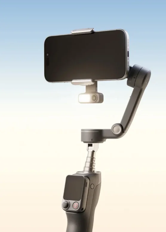Mavi ve bej tonlu arka plan önünde, küçük LED ışık aksesuarı takılmış elde taşınabilir stabilizer gimbal üzerindeki siyah bir akıllı telefon.