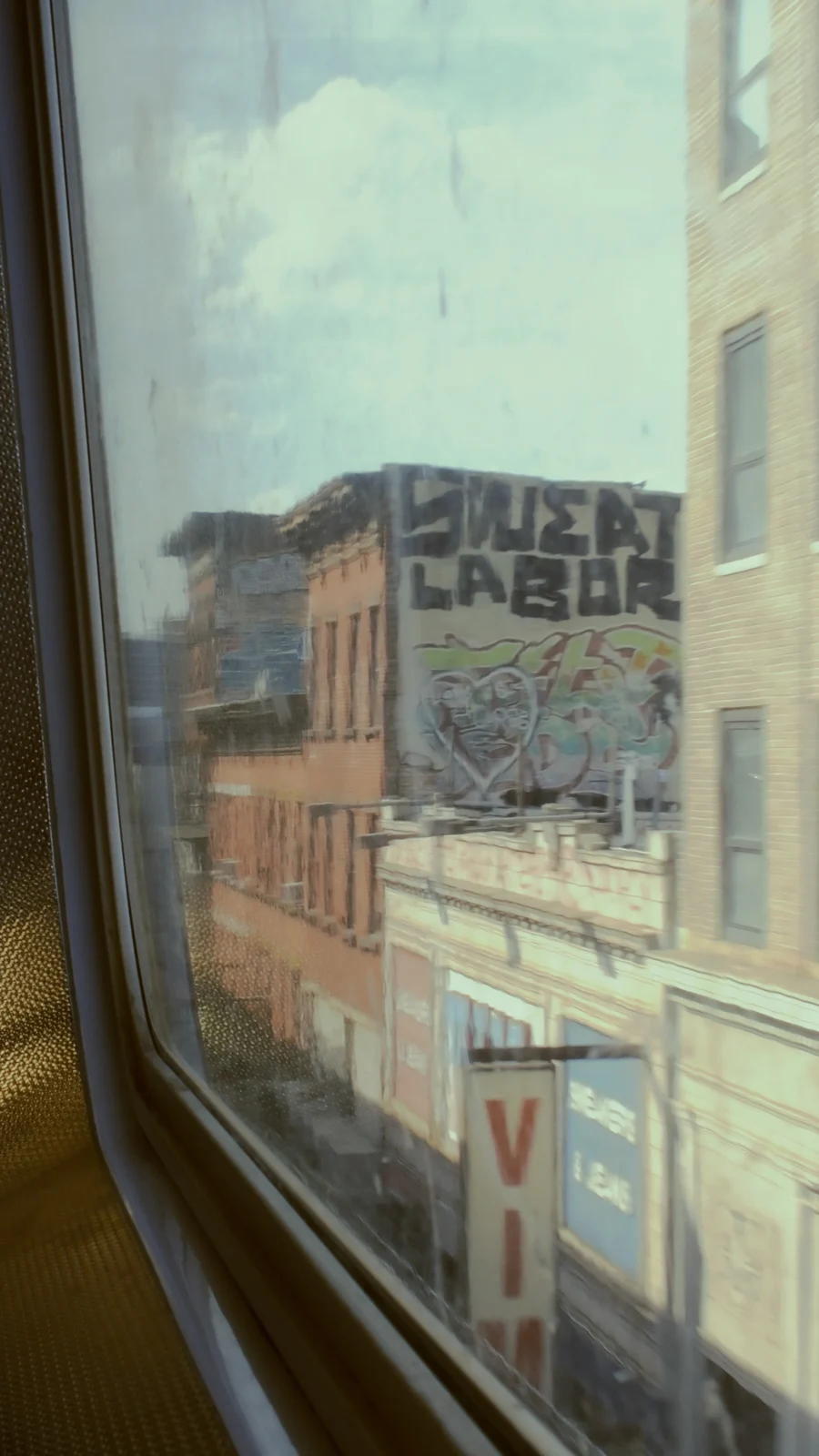 Kareden Önce: Bir Yönetmenin Sokak Fotoğrafçılığı Yaklaşımı 20 Bir pencerenin ardından görülen şehir sokağında tuğla binalar, duvarda “CHEAP LABOR” yazılı bir graffiti ve kısmen görünen “VIA” tabelası yer alıyor. Görüntü hafif bulanık.