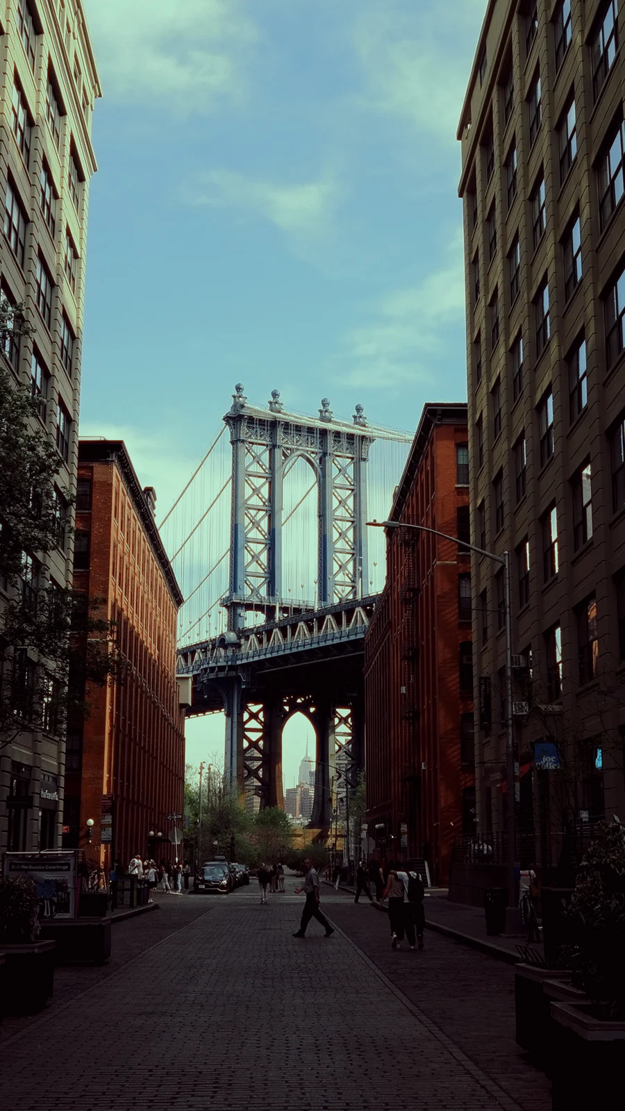 Kareden Önce: Bir Yönetmenin Sokak Fotoğrafçılığı Yaklaşımı 8 Brooklyn’de yüksek tuğla binaların arasından görülen Manhattan Bridge manzarası. Köprünün kemeri içinden Empire State Building seçiliyor. Kaldırım taşlı sokakta yayalar ve araçlar yer alıyor.