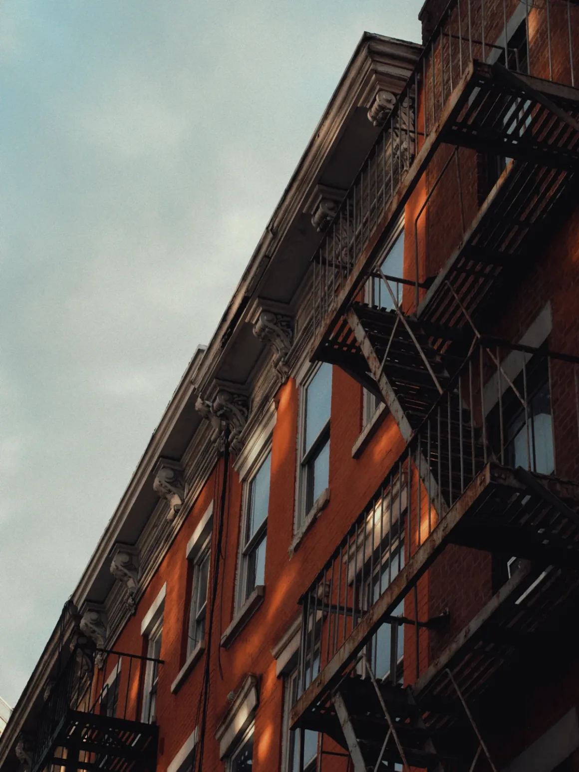 Kareden Önce: Bir Yönetmenin Sokak Fotoğrafçılığı Yaklaşımı 7 Siyah metal yangın merdivenlerine ve beyaz detaylara sahip tuğla apartman binası, parçalı bulutlu gökyüzü altında sıcak öğleden sonra ışığında aşağıdan yukarı doğru açıyla fotoğraflanmış.