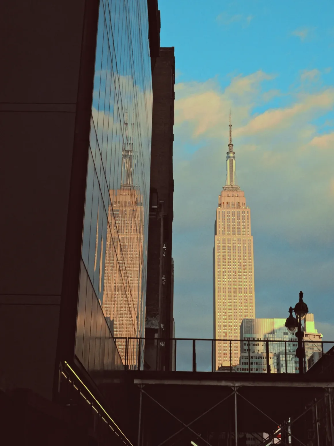 Kareden Önce: Bir Yönetmenin Sokak Fotoğrafçılığı Yaklaşımı 13 Empire State Building mavi gökyüzü ve bulutlar önünde yükseliyor. Ön plandaki cam yüzeylerde binanın yansıması görülüyor.