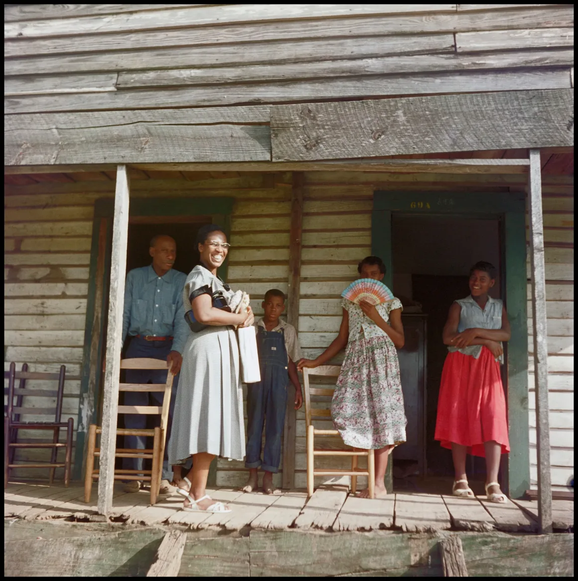 Beş kişi rustik bir ahşap verandada duruyor ve oturuyor. Bir kadın kameraya gülümsüyor; çocukların da bulunduğu diğer kişiler ise çevrede duruyor ya da oturuyor. Evin dış cephesi yıpranmış ahşap kaplamalar ve sade mobilyalarla dikkat çekiyor.