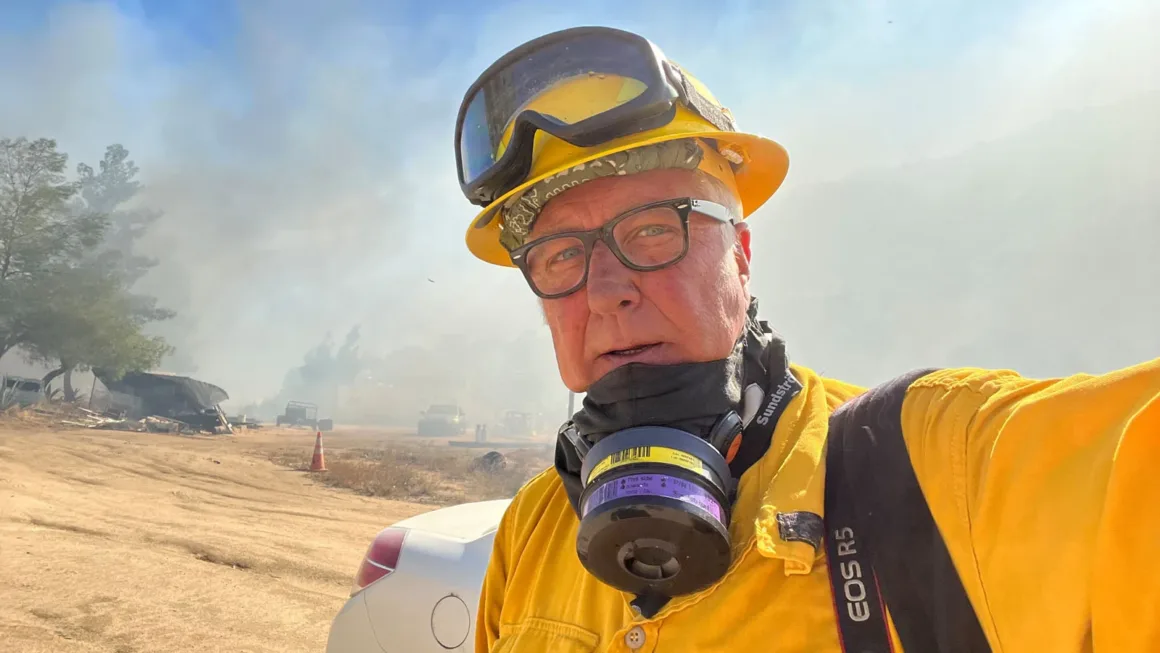 Pulitzer Ödüllü Foto Muhabiri Kanser Tedavisinin Ardından Yeniden Sahaya Döndü 2 Sarı koruyucu ekipman ve kask giyen bir itfaiyeci, dumanlı açık alanda selfie çekiyor. Arka planda yangın ekipmanları ve araçlar görülüyor.