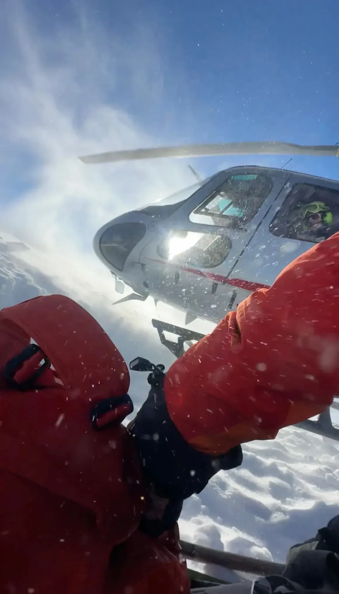 Bir helikopter karla kaplı zeminin üzerinde süzülürken, parlak kırmızı kış ekipmanı giyen bir kişi yoğun kar altında ilerliyor.