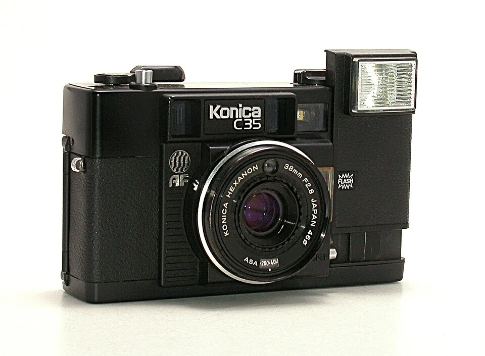 38mm lens ve dahili flaşa sahip siyah Konica C35 film kamerası, beyaz bir yüzey üzerinde hafif sola dönük şekilde konumlandırılmış. Kamerada görünür kadranlar ve dokulu tutma detayları bulunuyor.