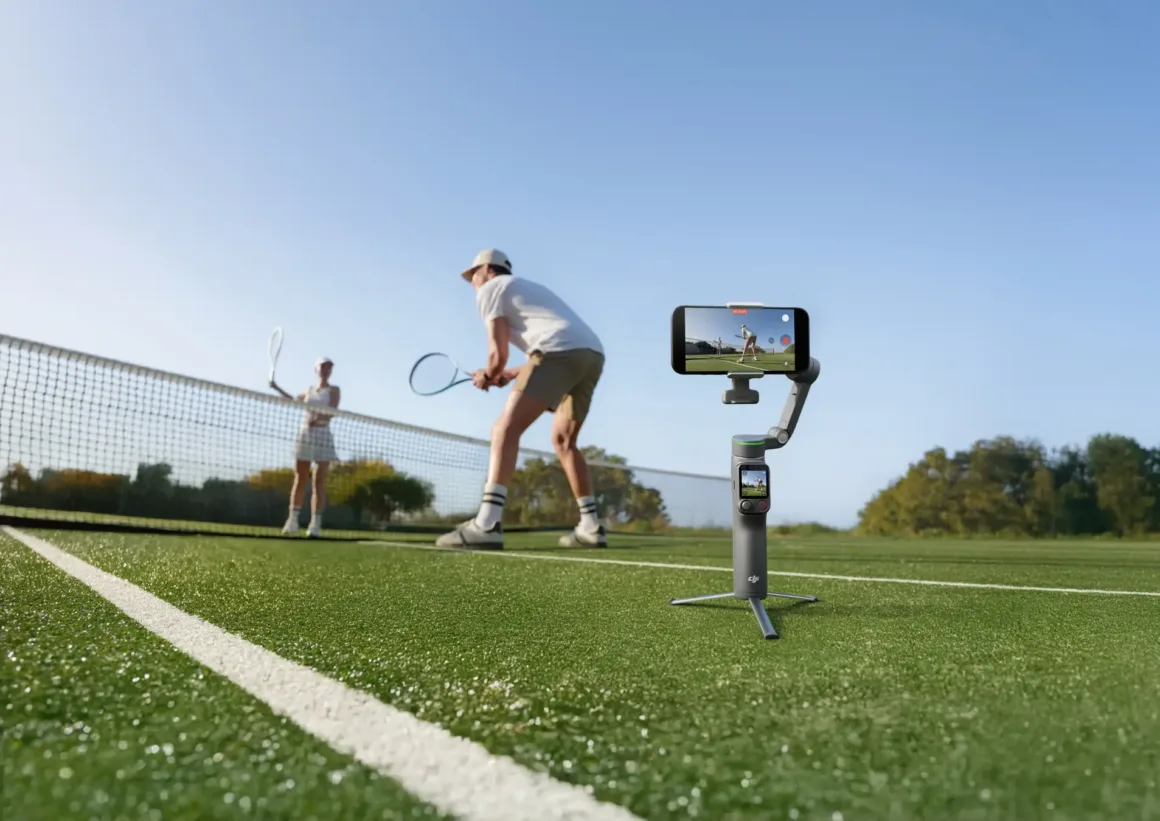 İki kişi açık hava tenis kortunda tenis oynarken, kortun kenarındaki tripodlu gimbal üzerine yerleştirilmiş akıllı telefon onları kaydediyor. Gökyüzü açık ve arka planda ağaçlar bulunuyor.