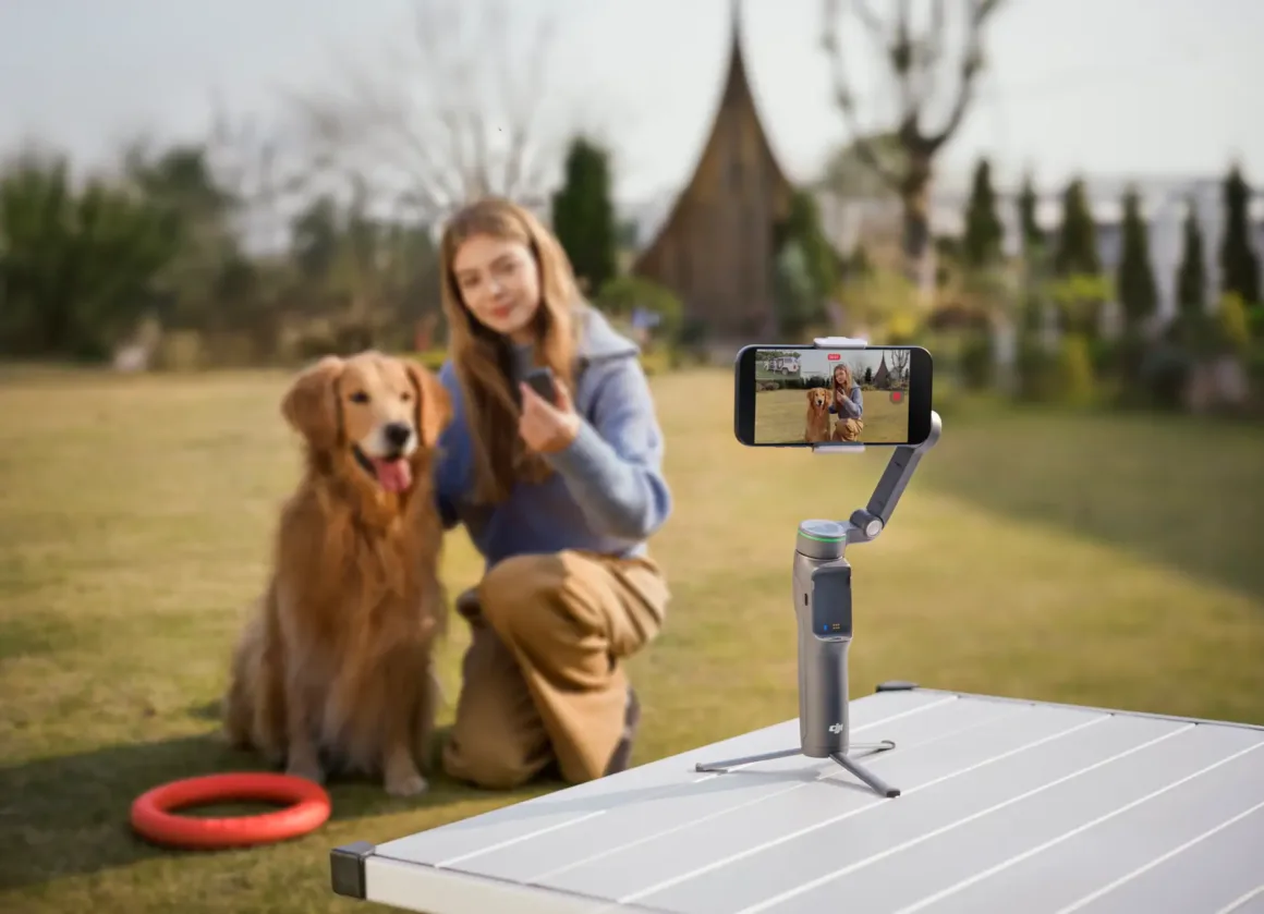 Genç bir kadın, çimlerin üzerinde golden retriever köpeğinin yanında diz çökmüş halde tripod üzerindeki akıllı telefonla selfie çekiyor. Yakında kırmızı bir frisbee bulunuyor; arka planda ise ağaçlar ve ahşap bir yapı yer alıyor.