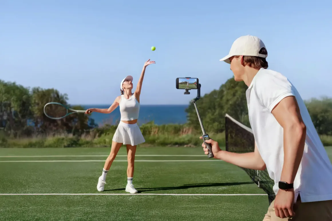 Beyaz tenis kıyafeti giyen bir kadın açık hava tenis kortunda servis atarken, beyaz giyimli bir erkek gimbal üzerindeki akıllı telefonla çekim yapıyor. Arka planda ağaçlar ve deniz görülüyor.