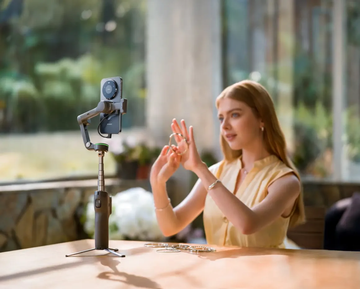 Bir kadın, tripod üzerine yerleştirilmiş akıllı telefonla kendisini kaydederken ellerindeki takıları gösteriyor. Büyük pencerelerden gelen güneş ışığı sahneye aydınlık bir atmosfer katıyor.