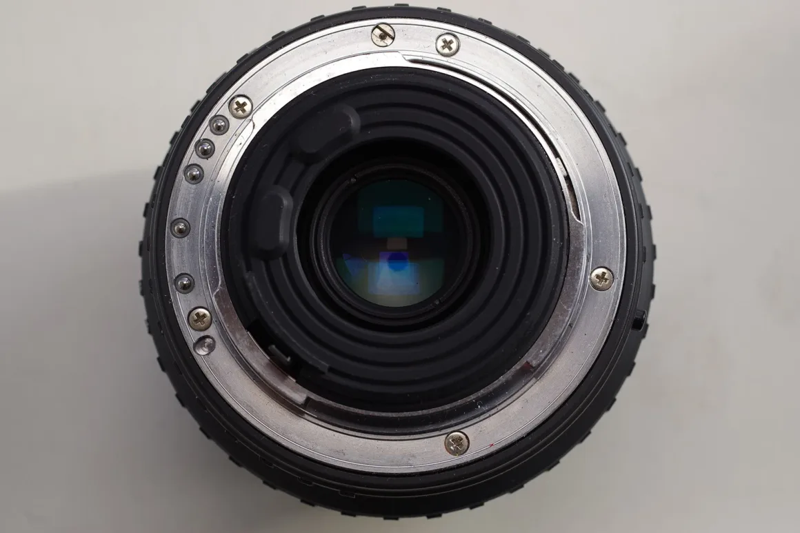 Düz açık renk arka plan önünde bir kamera lensinin arka tarafını gösteren yakın plan görüntü; metal lens yuvası, cam elemanlar ve montaj vidaları görülüyor.