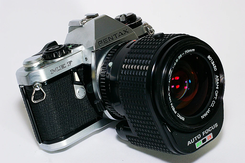 Beyaz bir yüzey üzerinde duran, büyük siyah otomatik netleme lensi takılı vintage Pentax ME F film kamerasının yakın plan görüntüsü.