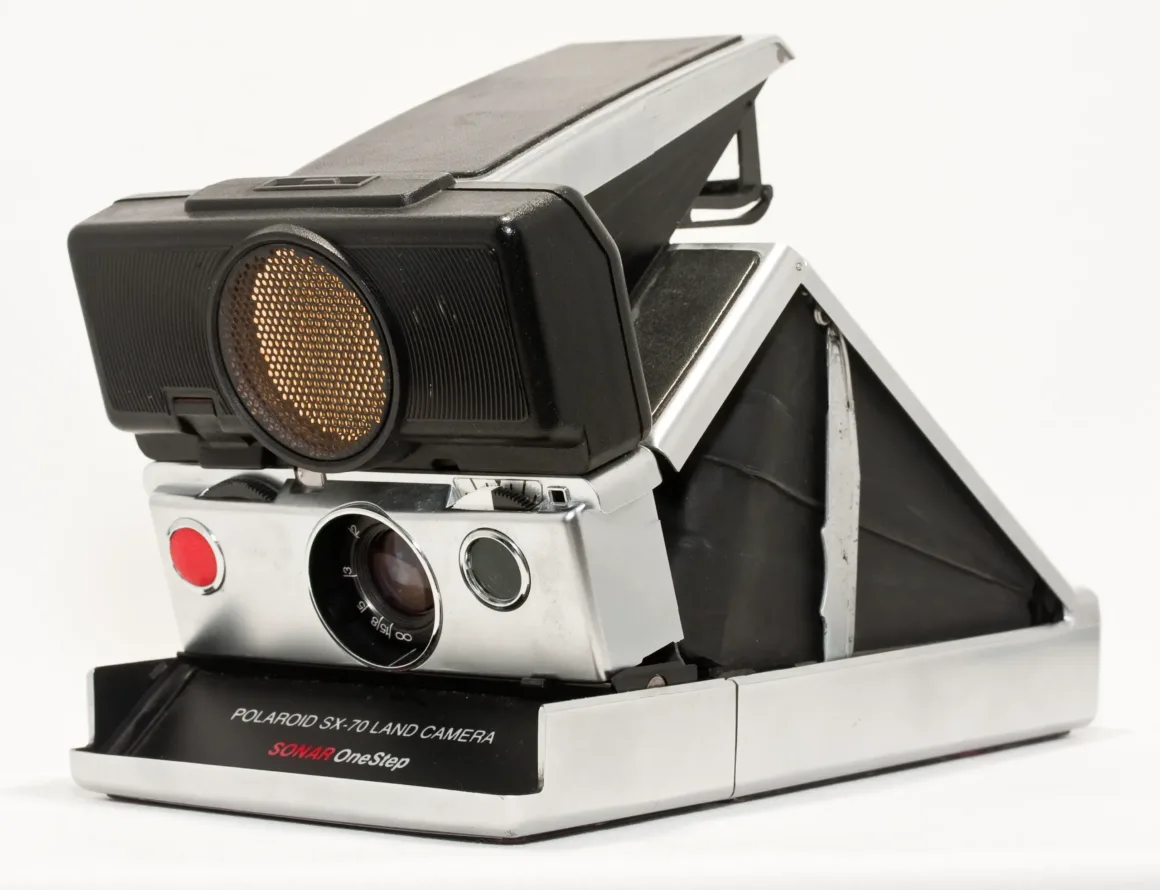 Beyaz arka plan önünde açık ve kullanıma hazır pozisyonda duran, lensi ve vizörü görünen gümüş-siyah vintage Polaroid SX-70 Land Camera.