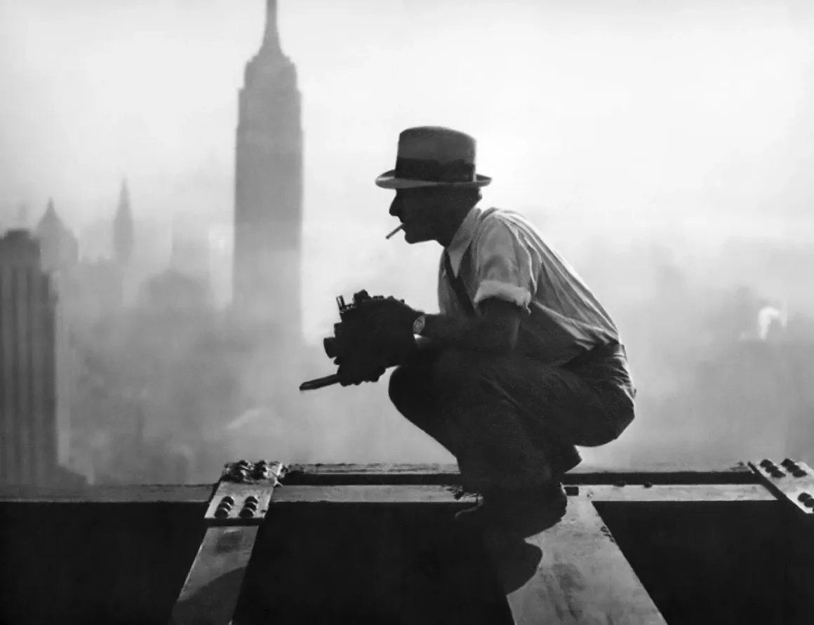 Şapka takan ve sigara içen bir adam, şehrin üzerinde yükselen çelik bir kiriş üzerinde çömelmiş durumda. Arka planda puslu Empire State Binası görülüyor. Elindeki kamera onun bir fotoğrafçı olduğunu düşündürüyor.