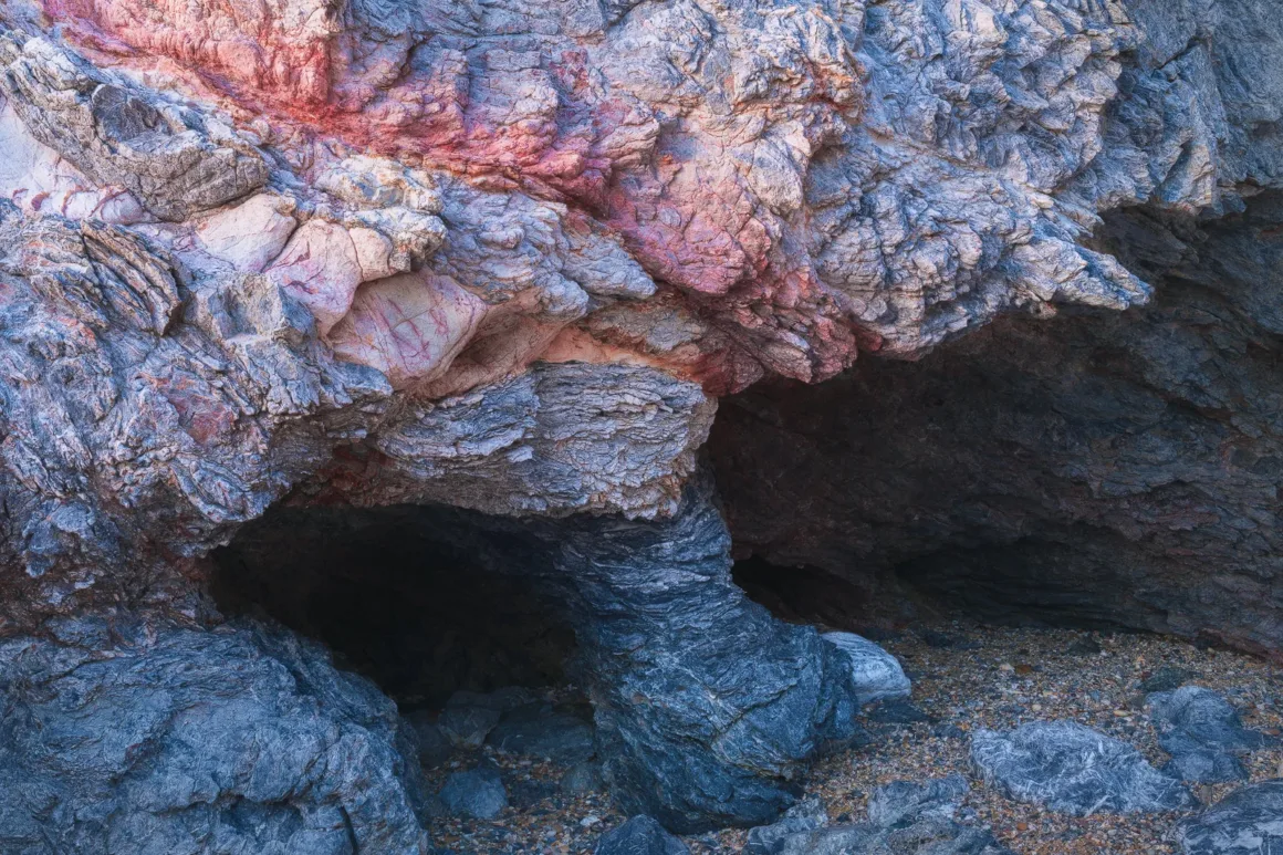Mavi ve gri tonlarında katmanlı, dokulu yüzeylere sahip kaya oluşumu; kırmızı ve pembe damarlar içeriyor. Alt kısımda küçük, karanlık bir mağara benzeri açıklık ve etrafında gevşek taşlar bulunuyor.