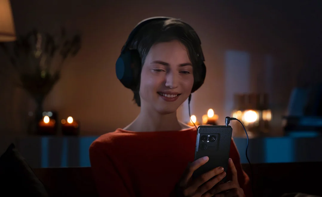 Kulaklık takan bir kadın loş ışıklı bir odada akıllı telefonuna bakarak gülümsüyor. Arka planda mum ışıkları yumuşak biçimde parlıyor.