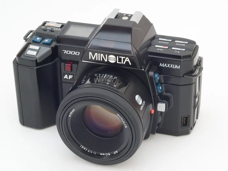 50mm lens takılı, çeşitli düğme ve kadranlara sahip siyah Minolta Maxxum 7000 otomatik netlemeli film kamerası beyaz arka plan üzerinde sergileniyor.