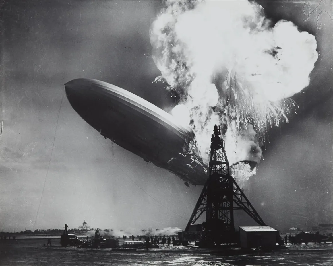 Hindenburg Felaketinin Fotoğrafları 89 Yıl Sonra Hâlâ Etkileyici GörünüyorDev bir hava gemisi, yüksek bir bağlama direğinin yakınında yere çakılırken alevler içinde kalıyor; gökyüzünü duman ve ateş kaplarken yerdeki insanlar olayı izliyor.