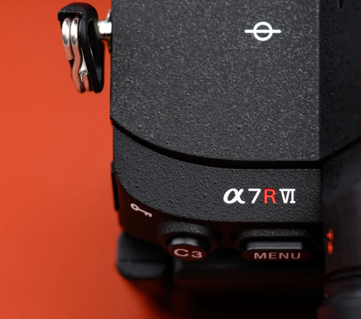 Turuncu arka plan önünde siyah Sony Alpha 7R VI kamera gövdesinin köşesine yakın çekim. Üzerinde model adı ve C3 ile MENU tuşları görünüyor.